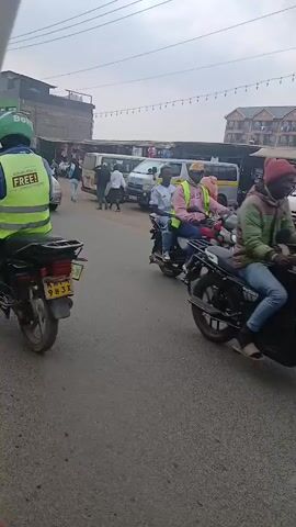 A motorcycle rider is seen in the foreground of the video, wearing a reflective vest and a helmet. The motorcycle is green and black. The rider is moving from left to right across the frame. In the background, there is a street with several vehicles, including vans, cars, and a motorcycle. There are also market stalls and buildings visible. People are walking and standing around the market area. The sky is overcast.