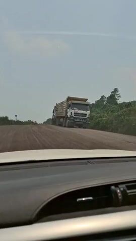 The video is shot from the interior of a vehicle, showing a dusty dirt road ahead. A procession of large dump trucks is visible on the road, moving in the same direction as the vehicle. The trucks are white and have large cargo beds. The road appears to be under construction or is unpaved. Vegetation is visible on the right side of the road and in the distance. The sky is overcast. The video pans slightly as the vehicle moves forward, revealing more trucks in the convoy.