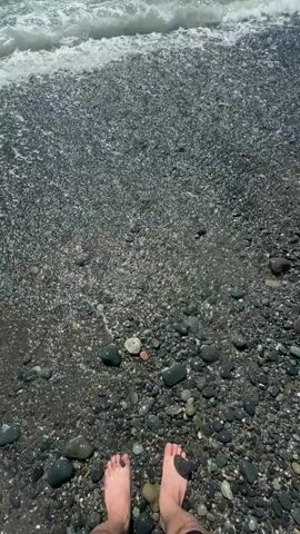 A first-person perspective video shows a person standing on a pebble beach with their feet bare. Waves are washing over the pebbles, coming up to the person's ankles. The water is clear and the pebbles are dark and varied in size. The sky is not visible, but the lighting suggests it is daytime.