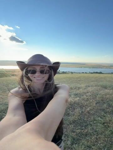 A woman in a cowboy hat and sunglasses is taking a selfie outdoors. She is smiling and looking at the camera. The background is a scenic landscape with a body of water and hills. The sun is shining and there are some clouds in the sky. The woman is wearing a black tank top and light-colored pants. She has long brown hair. She is standing on a grassy hill.
