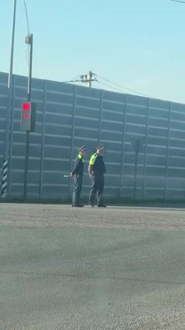 Two uniformed police officers are standing on the side of a road next to a traffic light that is red. One officer is holding a white stick. Behind them is a large, gray, soundproof barrier. The sky is blue.