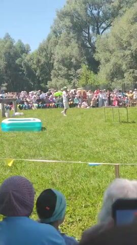 A wide shot shows a park with a crowd of people gathered on a grassy area. A person in a white outfit and green hat is on the grass in front of a low table. To the left is a blue inflatable pool. The person is running and seems to be performing. The crowd is seated and standing, with many people holding umbrellas. Trees and a blue sky are in the background. The video is focused on the performance.