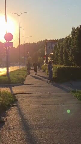 A group of five individuals are walking and jogging along a paved sidewalk. The sun is setting, casting a warm, golden glow over the scene and creating long shadows. A red car is seen driving on the road to the left of the sidewalk. Streetlights line the sidewalk, and trees and buildings are visible in the background. The overall atmosphere is peaceful and serene.