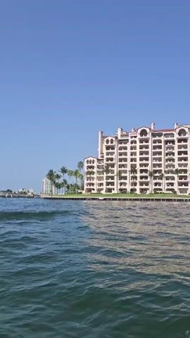A video recorded from a boat shows two identical, large, white apartment buildings with arched balconies and red tiled roofs situated on the shore. The buildings are surrounded by palm trees and a lush green lawn. The water in the foreground is blue and choppy, with white foam visible on the right side of the frame, indicating the boat is moving. The sky is clear and blue.