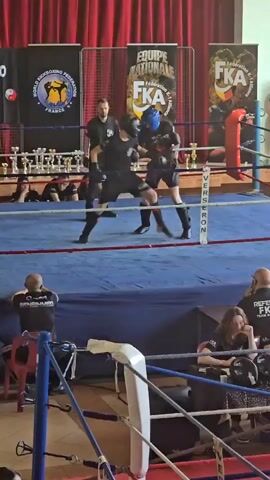 In a medium shot, two fighters compete in a kickboxing match inside a boxing ring with a blue canvas. The fighter on the right wears a black shirt with the number "8" on the back and black shorts. The fighter on the left wears a blue headgear, black shirt, and black shorts. They are exchanging kicks and punches under the supervision of a referee dressed in black.

The ring is surrounded by white ropes and blue padding. Behind the ring are red curtains and banners with logos of various kickboxing organizations. Trophies are displayed on a table behind the ring. Several spectators are seated around the ring, including some referees and coaches, all dressed in black shirts with logos.

The scene is brightly lit, and the overall atmosphere appears to be that of an amateur sporting event.