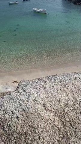The video shows a view from a large rock overlooking a calm, clear, turquoise body of water. There is a sandy beach at the edge of the water, and several small boats are anchored in the bay. The boats are white with some red accents. Rocky islands covered in green vegetation are visible in the background, along with a distant horizon under a clear blue sky. The sun is shining, creating ripples and light patterns on the water's surface. The camera appears to be stationary, offering a fixed perspective of the coastal scene.