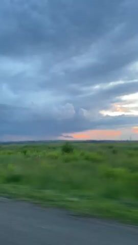 The video captures a scenic view from a motorcycle traveling along a road. The primary subject is the open landscape, characterized by vast green fields that blur due to motion. A prominent feature is the dramatic sky filled with heavy, dark clouds, hinting at impending rain or a storm. However, on the horizon, there's a sliver of warm, orange light from the setting sun peeking through, creating a striking contrast between the stormy sky and the calm sunset.

The perspective is from the rider, showing a hand in a black glove with pink lettering on the sleeve. The lettering is partially obscured but appears to be stylized text, possibly a brand or a personal emblem. A motorcycle mirror reflects a distorted image of the rider and the surrounding scenery, adding a personal touch to the video. The road surface is visible in the foreground, appearing to be paved with gravel or asphalt.

The time of day is clearly late afternoon or early evening, as indicated by the sunset. The weather appears turbulent, with dark clouds suggesting a storm or heavy rain. The location, Старое Мартьяново, Russia, suggests a rural or semi-rural setting, consistent with the open fields and expansive sky. The overall mood is one of adventurous travel, appreciating the raw beauty of nature, and perhaps a touch of melancholy or awe inspired by the dramatic weather. There are no other people or distinct objects visible besides the motorcycle parts and the landscape.