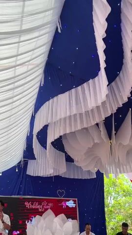 The video captures the interior of a meticulously decorated event tent, likely set up for a wedding or celebration. The ceiling is predominantly covered in a deep blue fabric, dotted with numerous small, twinkling blue and warm white LED lights, creating an illusion of a starry sky. From this blue backdrop, an array of elaborate white fabric decorations hangs down. These include cascading, ruffled white fabric strips arranged in elegant curves, and multi-layered, fan-like white fabric structures that add depth and texture. Sheer white fabric is also draped along the side walls of the tent in gentle swags.

At the bottom of the frame, a digital display or screen is visible, featuring a red and black background with decorative elements. The text "Wedding day" is clearly displayed on this screen, indicating the specific nature of the event. Upbeat Vietnamese music plays throughout the video, contributing to a lively and celebratory atmosphere. The overall aesthetic, with its blue and white color scheme, sparkling lights, and decorative fabric, evokes a festive and romantic ambiance. No individuals are visible in the shot, but the setting is clearly prepared for a special occasion.