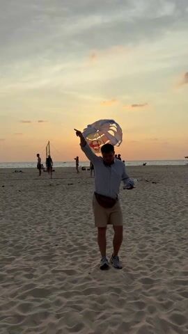 The video shows a man standing on a sandy beach, holding a bottle of water. He is wearing a light blue shirt, tan shorts, and gray sneakers. Behind him, people are playing volleyball, and further back, a parasail is in the air against the backdrop of a sunset. The sky is a mix of orange and gray.