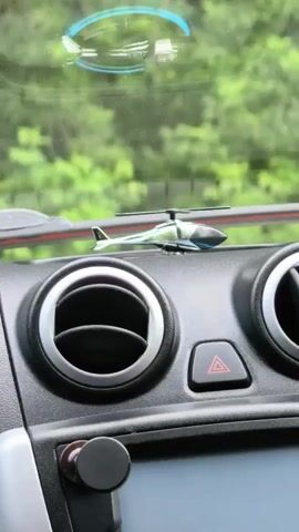 The video shows a close-up view of a car's dashboard. In the center of the dashboard, a small toy helicopter is placed on top of the air vents. To the left and right of the helicopter are two circular air vents with chrome trim. Between the vents and slightly behind the helicopter is a red hazard light button. Below the vents, a portion of the car's infotainment screen is visible. A black circular object, possibly a phone mount, is attached to the dashboard below the left air vent. The background visible through the windshield is blurred, showing green foliage, suggesting the car is in motion outdoors. A reflection of the toy helicopter, appearing larger and slightly distorted, is visible on the windshield above the dashboard. The camera angle is slightly upward, focusing on the dashboard and the toy helicopter.