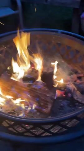 The video shows a close-up of a fire pit with logs burning. Sparks fly upward from the flames. The fire pit is round with a decorative metal grate. The flames are bright orange and yellow, illuminating the surrounding area with a warm glow. The background is dark, suggesting it is nighttime or dusk. The video captures the dynamic movement of the fire, with flames licking and dancing around the wood.