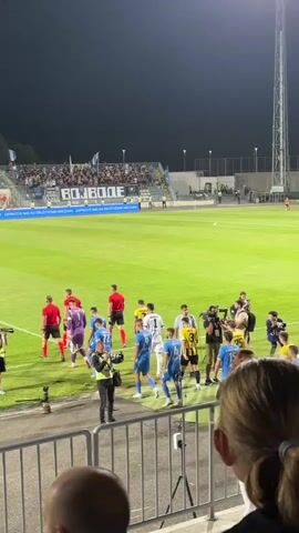 The video shows a football match at night. Players in blue and yellow uniforms are walking on the field. Spectators are in the stands, and some are holding banners. There are also cameras and crew members filming the event. The scene appears to be at the end of a match, as players are interacting with each other and the crew.