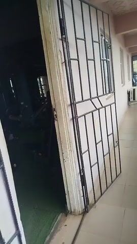 The video opens to a view of a gym entrance, with a metal security gate partially blocking the view into the interior. The gate has a grid pattern of vertical and horizontal bars, with a small window near the top. As the camera moves forward and the gate opens, the interior of the gym becomes visible. The gym appears to be dimly lit with a dark ceiling and light gray walls. The floor is covered in green matting. Several pieces of exercise equipment are visible, including weight benches, barbells with weights, and what appears to be a weight machine in the foreground. A person in a dark shirt and white towel around their neck is seated on a piece of equipment in the middle ground, facing away from the camera. Further in the background, another person in a white shirt is standing near a weight machine, and another person in a light blue shirt is partially visible. The overall impression is of a functional, if somewhat basic, gym environment.