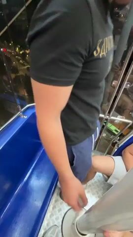 From inside a Ferris wheel car at night, a man wearing a black t-shirt and a fedora is visible, looking out at the illuminated city below. The car has blue seating and a glass barrier with metal framing. The sky is dark, with the outline of a mountain range visible in the distance. The inside of the Ferris wheel car is illuminated with purple and blue lights. There are other people in the car as well.