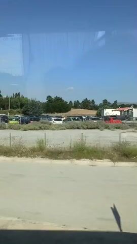 The video shows a view from inside a vehicle looking out at a parking lot on a sunny day. The parking lot is filled with various cars and some bushes and trees are visible in the background. A utility pole with a sign is also present.