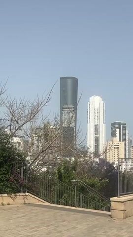 The video shows a view of a city skyline, featuring several high-rise buildings of varying designs and colors. In the foreground, there's a paved area with a metal railing, and some trees and bushes, including some with purple flowers. The sky is light blue and clear. The camera slowly pans from left to right, revealing more of the cityscape. There are no people visible in the frame.