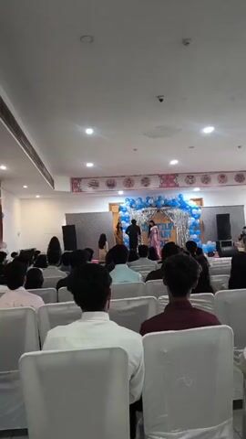 The video shows an indoor event, likely a ceremony or performance, taking place in a hall. In the foreground, the backs of several rows of white chairs are visible, occupied by an audience facing a stage. The audience members appear to be young adults, dressed in formal attire, with men in shirts and women in dresses. On the stage, a small group of people are present, some standing and some possibly performing. The stage is decorated with blue and silver balloons, and there are speakers on either side. The ceiling is white with recessed lighting, and a large banner with decorative circular images hangs above the stage. The overall atmosphere seems to be one of a festive gathering.