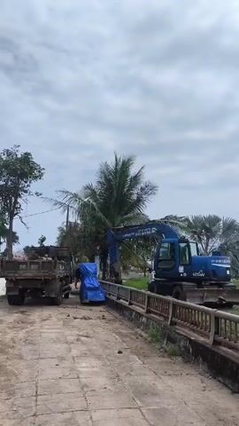The video shows a blue Hitachi excavator filling the back of a dump truck with dirt.  The excavator is parked on a brick bridge with a railing and green vegetation behind it. The dump truck is an older model, and there is a blue tarp covering something on the road near the truck.  Several palm trees are visible in the background, and the sky is overcast. The area appears to be in a tropical climate.  Music is playing in the background.