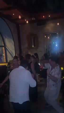 The video shows a dimly lit indoor space, possibly a bar or restaurant, with people dancing. In the foreground, two men are dancing. The man on the left wears a white button-down shirt and dark pants. The man on the right wears a floral-patterned short-sleeved shirt and light-colored pants. Several other people are visible in the background, some dancing and some standing. The environment features exposed wooden beams on the ceiling, decorative lighting fixtures, and a large arched window on the left side of the frame, through which some external lights are visible. There are mirrors on the wall in the background. The overall atmosphere appears to be lively and social.