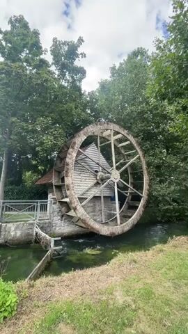 A water wheel is turning next to a stone and wooden building, surrounded by trees and a flowing river. The sky is cloudy.