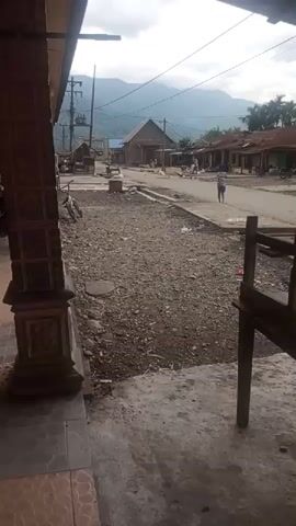 A casual video shows a residential street lined with small buildings. A person walks away from the camera towards a larger building in the distance, which appears to be a church or community hall. Several other figures, including children, are visible in the background, engaged in activities typical of a village setting. The street is paved in some areas and covered with gravel in others. Mountains loom in the background under a cloudy sky. A bicycle is parked near the left side of the frame. The overall impression is one of a peaceful, rural community.