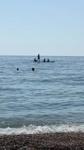 The video shows a wide shot of a calm sea under a clear blue sky. In the foreground, a pebble beach is visible, with small waves gently breaking on the shore. Further out in the water, several people are enjoying recreational activities. In the mid-ground, three individuals are swimming, with their heads above the water. In the background, a group of five people are on a paddleboard. One person is standing at the back of the paddleboard, appearing to steer or balance, while the other four are seated or kneeling on the board. The overall impression is a peaceful day at the beach with various water activities taking place.