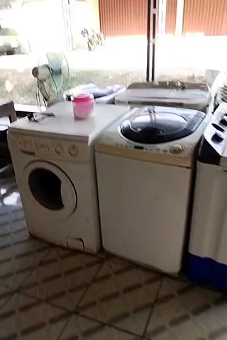 The video shows a row of washing machines outside, with a table and chair in the foreground. The washing machines are of various types and brands. In the background, there is a vehicle and some buildings. The scene appears to be a commercial area, possibly a laundromat or a shop selling used appliances.