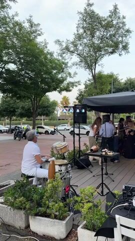 The video shows an outdoor live music performance in front of a building. A band is set up on a wooden platform under a black canopy, playing for a small crowd. The band includes a conga player, a drummer, and several other musicians.  The crowd is standing and mingling, some people are dancing. The area is paved with red bricks and there are trees and potted plants around. Cars are parked in the background. The video pans right to show more of the crowd and building.