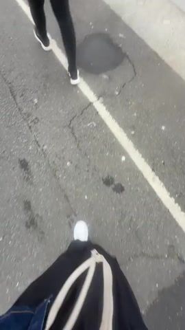 A first-person perspective video shows two people walking along a paved path next to a body of water. The path is cracked and has a white line marking its edge. In the foreground, one person's legs are visible, wearing black pants and white sneakers, carrying a light-colored bag. Behind them, the legs of another person are visible, also wearing black pants and black and white sneakers. The path leads to a docked white ferry named "FORTUNA" with a blue hull. The ferry is tied to the dock with thick ropes. There are orange and white traffic cones on the dock near the ferry. The sky is overcast.