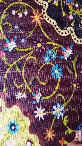 A close-up video shows a textured mat with a dark brown background and a floral design featuring blue, white, pink, orange, and yellow flowers. Green swirling vines connect the floral elements. The mat has a textured surface with a grid-like pattern. A hand briefly enters the frame at the bottom right corner. The camera zooms slightly inward, focusing on a section with blue flowers and green vines.