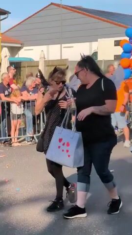 A parade is underway on a street in front of a yellow building. Two women walk side-by-side, one wearing a black dress with white polka dots and devil horns, the other in a black t-shirt and blue jeans. Both women are holding drinks. A large blue tote bag with red hearts is carried by the woman in the polka dot dress. A man wearing an orange shirt and blue shorts is seen near a truck adorned with balloons. The truck has the words "Dorfclub Hardisleben e.V." and "LACKSERVICE Pfundheller" written on it. Other people, some with costumes, are visible in the background, watching the parade. The ground is covered with confetti.