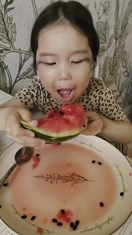 A young Asian girl with dark hair is seated at a table, joyfully consuming a large wedge of watermelon. She is wearing a leopard-print shirt and sits behind a white plate that is stained pink with watermelon juice and scattered seeds. A tarnished silver spoon lies on the plate. The girl smiles intermittently as she eats, looking to her left and then directly at the camera. The background is adorned with a floral-patterned wallpaper.
