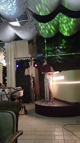 The video shows a woman standing on a circular stage in front of a DJ booth. She is wearing a long, white, sparkly dress with a high slit and appears to be singing or speaking into a microphone. There is a music stand in front of her with sheet music. The stage is lit with blue and white lights. Behind the DJ booth, there is a large screen displaying abstract patterns. The ceiling is decorated with undulating fabric that is lit with green and blue lights, creating a wavy, textured effect. To the left of the stage, there are speakers and some lighting equipment. In the foreground, to the left, a stroller and a portion of a couch are visible. The floor appears to be tiled. The overall environment suggests an indoor venue, possibly a restaurant or club, with a performance setup. The lighting and decorations create a festive and atmospheric mood.
