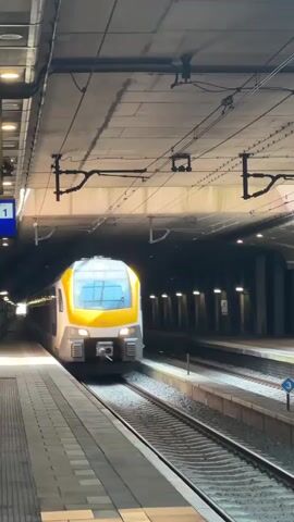 The video shows a train pulling into an underground station. The train is yellow and blue and has "Tilburg Universiteit" displayed on its front. The platform is tiled in a light brown color. There are overhead power lines and lights on the ceiling of the station. The walls of the tunnel are grey. The train is moving slowly as it approaches the platform.