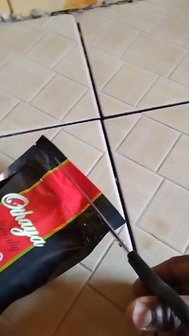 A hand uses scissors to cut open a bag of "Chibaya Specialty Coffee". The bag is black with a red and yellow label. It sits on a tiled surface with dark grout lines.