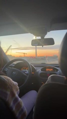 A person is driving a small car, an Opel, at sunset. The driver wears a striped sweater and light purple pants. The car's dashboard is visible, with a digital display showing "20:06" and "14C". The car is driving on a road with orange traffic cones on both sides. There is a crane and some buildings in the distance, silhouetted against the bright orange and yellow sky. The driver is talking to a passenger in the front seat, who is partially visible.