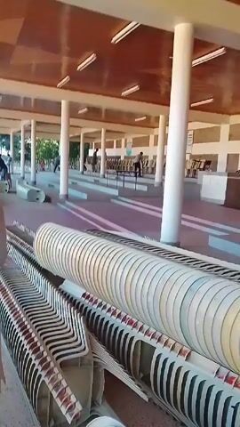 The video depicts the interior of a large, open-air building, likely a pavilion or covered area, supported by white columns. The ceiling is made of brown wooden paneling with evenly spaced fluorescent lights. The floor has a series of long, parallel, light-colored platforms or lanes, separated by recessed areas.  A stack of white plastic chairs is prominently positioned in the foreground, taking up a significant portion of the bottom of the frame. Several people are visible in the background, walking around or standing. Some of them appear to be setting up rows of chairs at tables in the distance.  A woman wearing a light-colored coat walks into the frame from the left, moving away from the stack of chairs.  The overall impression is that the space is being prepared for an event.