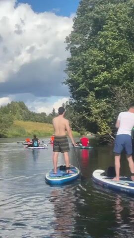 A group of people are paddleboarding on a river. The river is surrounded by trees and has a cloudy sky. There are at least four paddleboards visible, each with one or two people on it. The paddleboarders are moving in the same direction. Some are standing and paddling, while others are sitting.