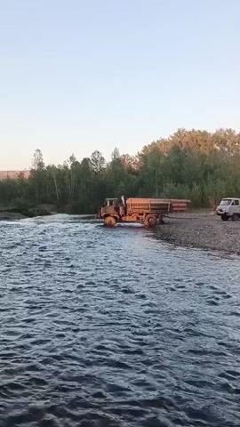 A large, orange truck is partially submerged in a shallow, rocky river, its front wheels turned towards the far bank. The truck is loaded with long, light-colored cylindrical objects, likely logs. The river flows from left to right across the frame, with the water reaching the middle of the truck's tires. The truck is positioned on the right side of the river, near a gravelly bank. To the right of the truck, a smaller white truck is parked on the bank. The background consists of a dense line of green trees, with a clear, pale sky above. The overall scene depicts a challenging and potentially risky operation of transporting goods across a natural obstacle.