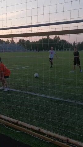 The video shows a soccer field with a goal in the foreground. Four young people are visible. Two are near the goal, one in an orange shirt and one in a black shirt. They are both hunched over and appear to be interacting with something on the ground. A soccer ball is on the ground near them. In the background, two more young people are visible on the field. One is wearing a white t-shirt and black shorts, and the other is wearing a black t-shirt and black shorts. They are standing further down the field. The sky in the background appears to be dusk or dawn with a pale pinkish hue. There are bleachers and trees visible in the distance.