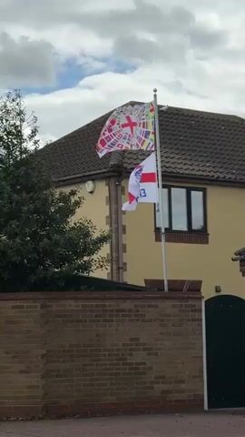Two flags are displayed on a flagpole in front of a two-story house with a brown tiled roof. The bottom flag is white with a red cross and a blue emblem. The word ENGLAND is written on it in blue lettering. The top flag is white with a red cross in the center, surrounded by a multitude of small national flags. A brick wall runs along the bottom of the frame. The sky is cloudy.
