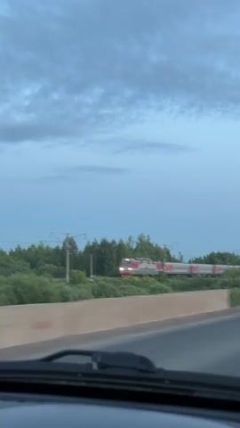 A video clip recorded from inside a moving vehicle captures a train passing by on a parallel track. The train, adorned with the red "РЖД" (Russian Railways) logo on its white and grey carriages, moves from right to left across the frame, partially obscured by green foliage. The scene is set outdoors, with a line of trees visible behind the train, separating the railway line from what appears to be a road or highway in the foreground. The sky above is a light blue with scattered clouds, suggesting either dusk or dawn. The view is partially framed by the top edge of a car's dashboard or windshield wipers at the bottom of the screen. The overall setting appears to be a rural or suburban area in Совхозный, Russia.