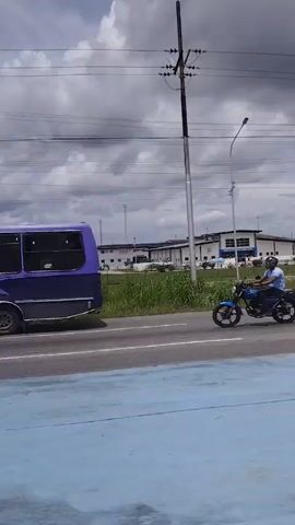 The video captures a cloudy sky over a landscape with a low-rise building in the background. A road runs in the foreground with a blue and yellow painted surface. Power lines and poles are visible against the sky. A line of grass and bushes separates the road from the building. Several vehicles, including a white truck, a red bus, and motorcycles, are parked in front of the building. The building appears to be an industrial or commercial facility with multiple sections and a sign above an entrance. The camera pans slightly to the right, revealing more of the surrounding environment, including distant hills.