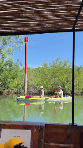The video shows two people in a yellow kayak paddling on a calm, green river. They are both wearing hats and long-sleeved shirts. The person in the front is wearing a light-colored hat and a tan shirt, and the person in the back is wearing a darker hat and a tan shirt. They are both using red paddles. In the background, there is a tall, red and white pole, and lush green trees line the riverbank. The sky is bright blue with a few white clouds. The video is shot from a perspective inside a wooden structure, possibly a boat or a covered area, with a thatched roof overhead. A portion of a yellow object and some text are visible in the foreground on the left.