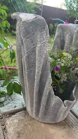 The video shows a close-up, slow-motion shot of a concrete planter decorated with a textured silver finish. The planter is shaped like a draped fabric and is filled with a variety of green plants, white flowers, and small purple and yellow blossoms. The planter sits outdoors on a surface that appears to be made of concrete slabs. In the background, there is lush green foliage, a pink garden hose, and a green metal fence. The lighting suggests it is daytime, possibly late afternoon due to the soft, natural light. The camera slowly pans around the planter, highlighting its unique shape and the flowers within.