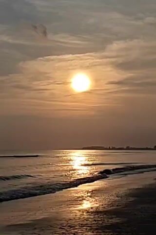 The video captures a scenic view of the sun setting over the ocean. The sun appears as a bright orb amidst a sky filled with wispy clouds, casting a golden hue across the scene. The water reflects the sunlight, creating a shimmering path towards the shore. Gentle waves lap against the sandy beach. In the distance, a faint outline of land or trees can be discerned along the horizon.