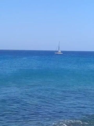 The video opens with a wide shot of a calm, deep blue ocean under a clear sky, with a white sailboat sailing from left to right in the middle distance. The foreground water is a lighter turquoise. As the sailboat disappears from view, the camera pans right and downwards, revealing a rugged rocky coastline. Waves are visible crashing against the dark, possibly volcanic, rocks that line the shore, creating white foam. The rocks in the foreground are dark and appear rounded. The clear blue sea stretches out to the horizon under a bright, sunny sky, indicating daytime conditions in Puerto Del Carmen, Spain.