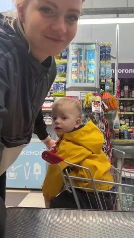 A young woman with blonde hair pulled back and wearing sunglasses on her head pushes a metal shopping cart. A young child wearing a yellow raincoat is sitting in the child seat of the cart. They are in a grocery store, near the checkout line. The child is smiling and holding onto the safety bar of the shopping cart. Another person wearing a burgundy vest walks behind the woman. Shelves stocked with various goods are visible in the background, along with a refrigerated display case filled with items.