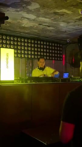 Here is a description of the video:
The video features a DJ behind a DJ booth in a dimly lit club or event venue. The DJ is a man wearing a yellow shirt and headphones around his neck. He is focused on the equipment in front of him, which includes a Pioneer DJ mixer and two laptops. He interacts with the mixer, adjusting levels and presumably mixing tracks.
Behind the DJ is a wall adorned with numerous circular lights arranged in a grid pattern. To the left of the DJ booth, a vertical sign or banner reads "bar américas" in white text.
The ceiling has a rough, textured appearance, possibly concrete. Red lighting elements are also visible, adding to the ambiance of the space. A fan can be seen in the background. The camera's perspective is from a slightly low angle, likely held by someone standing in the audience or nearby. The overall scene suggests a live music event or club setting where the DJ is performing.