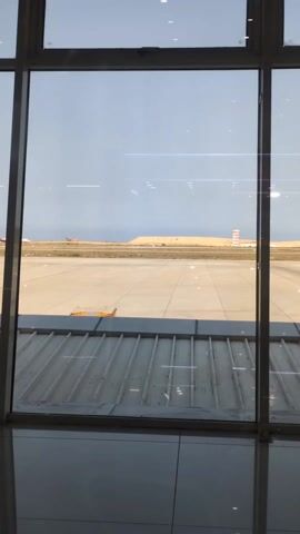 The video shows an airplane taxiing on a runway, as seen through a large window. The runway is bordered by sand dunes. In the background, there is a control tower visible. The window is segmented by a white frame, and a metal grating can be seen below the window. The sky is overcast.